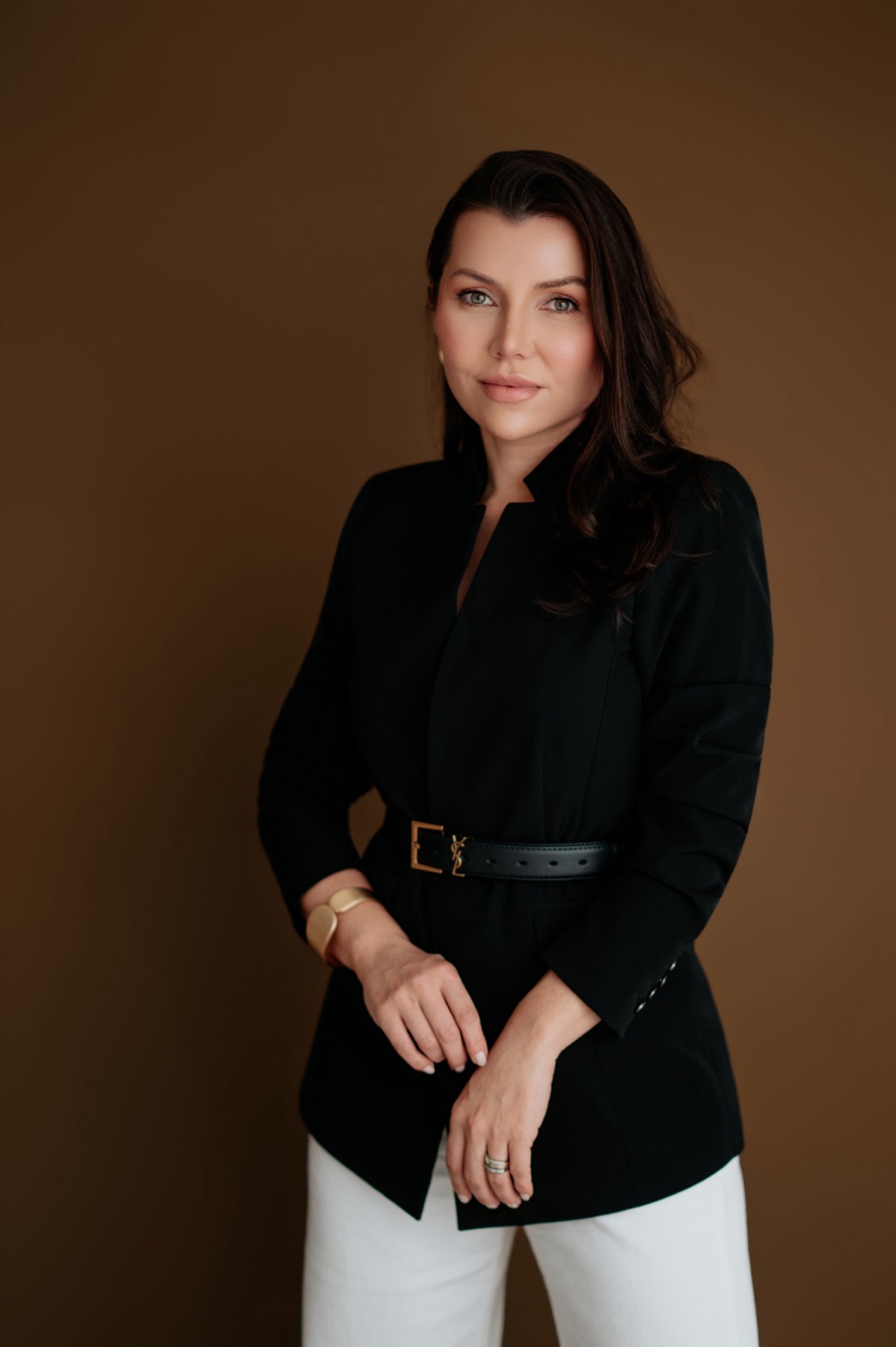 Dra. Letícia Melo em pé, de blazer preto, em fundo marrom